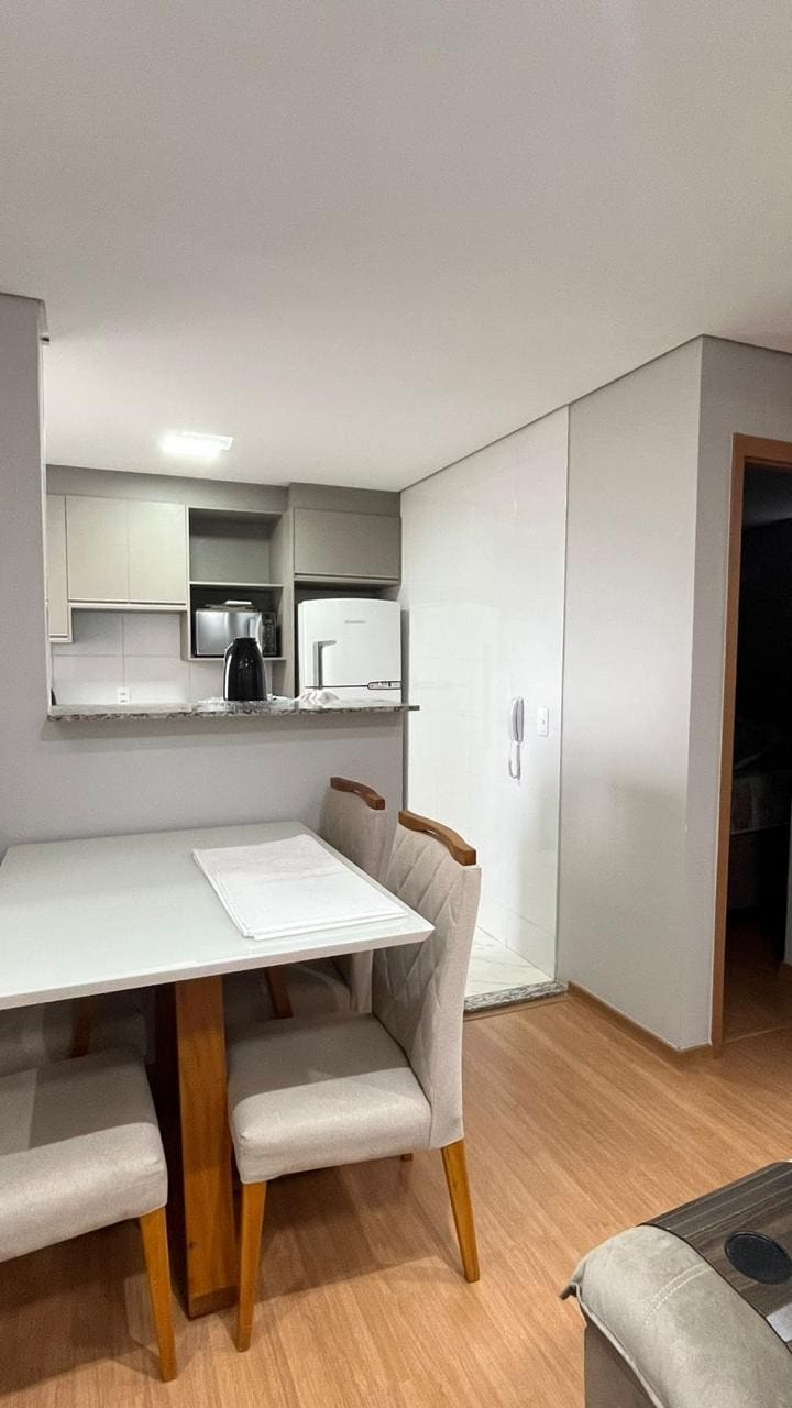 A modern dining area is shown, featuring a light-colored table with four upholstered chairs. The kitchen is visible in the background, equipped with sleek cabinets and appliances. Natural light brightens the space, enhancing the clean, minimalist aesthetic.