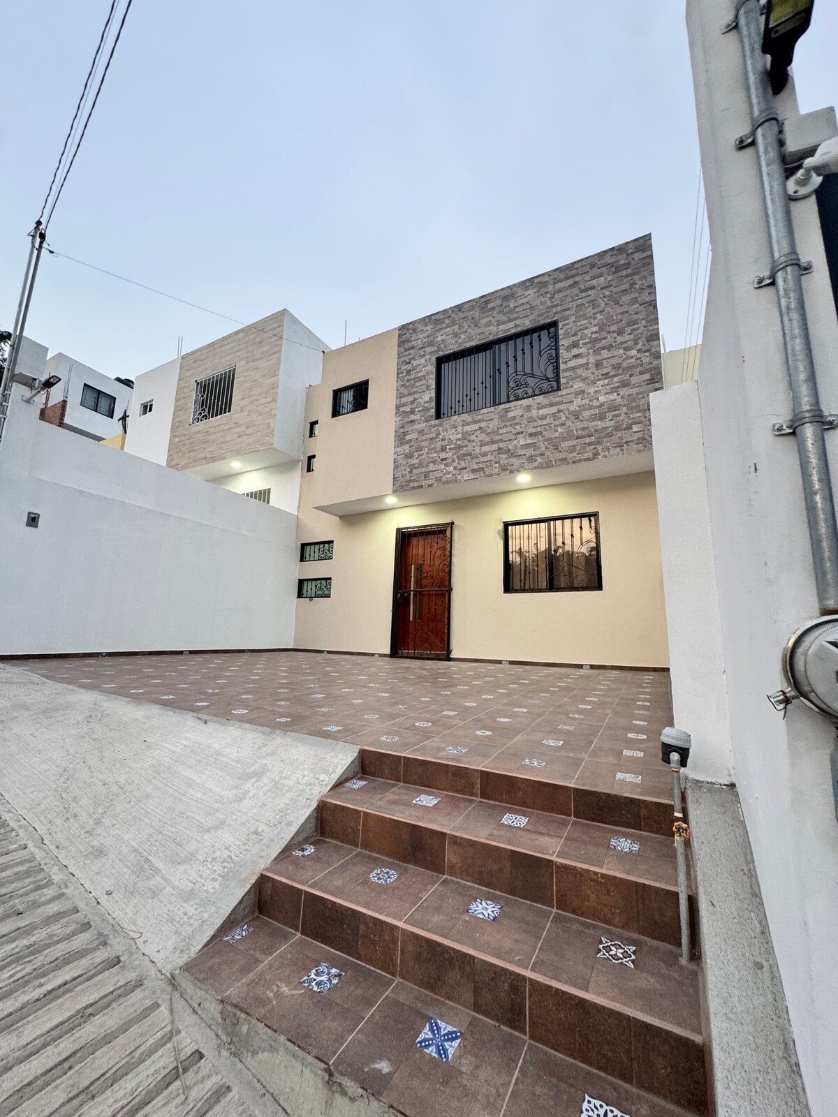 The exterior of the accommodation displays a modern two-story structure with a mix of light-colored walls and stone accents. A staircase leads to the inviting entrance, framed by a tiled patio that features a geometric pattern, providing space for outdoor relaxation.