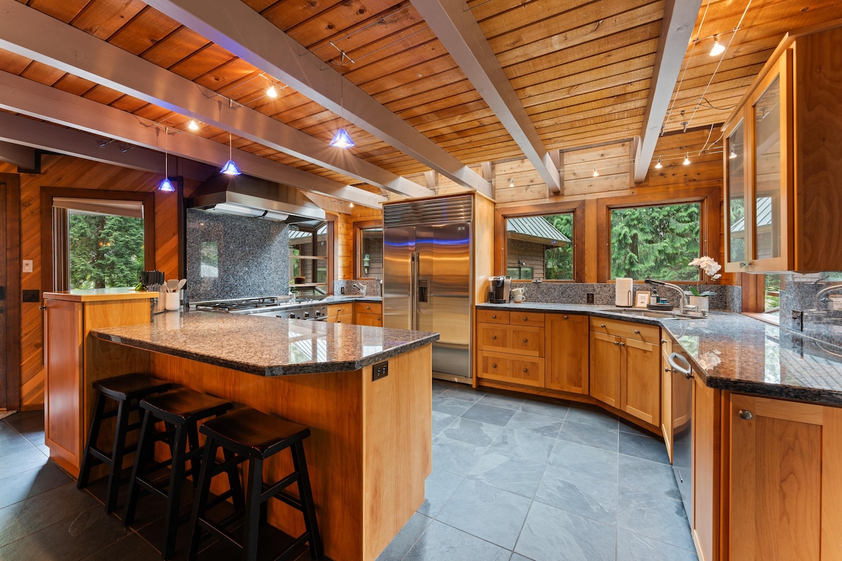 A spacious kitchen features wooden cabinetry and a large central island with bar seating. Natural light floods the room through large windows, highlighting the dark granite countertops and stainless steel appliances. A gas stove and ample counter space are provided for cooking and meal preparation.