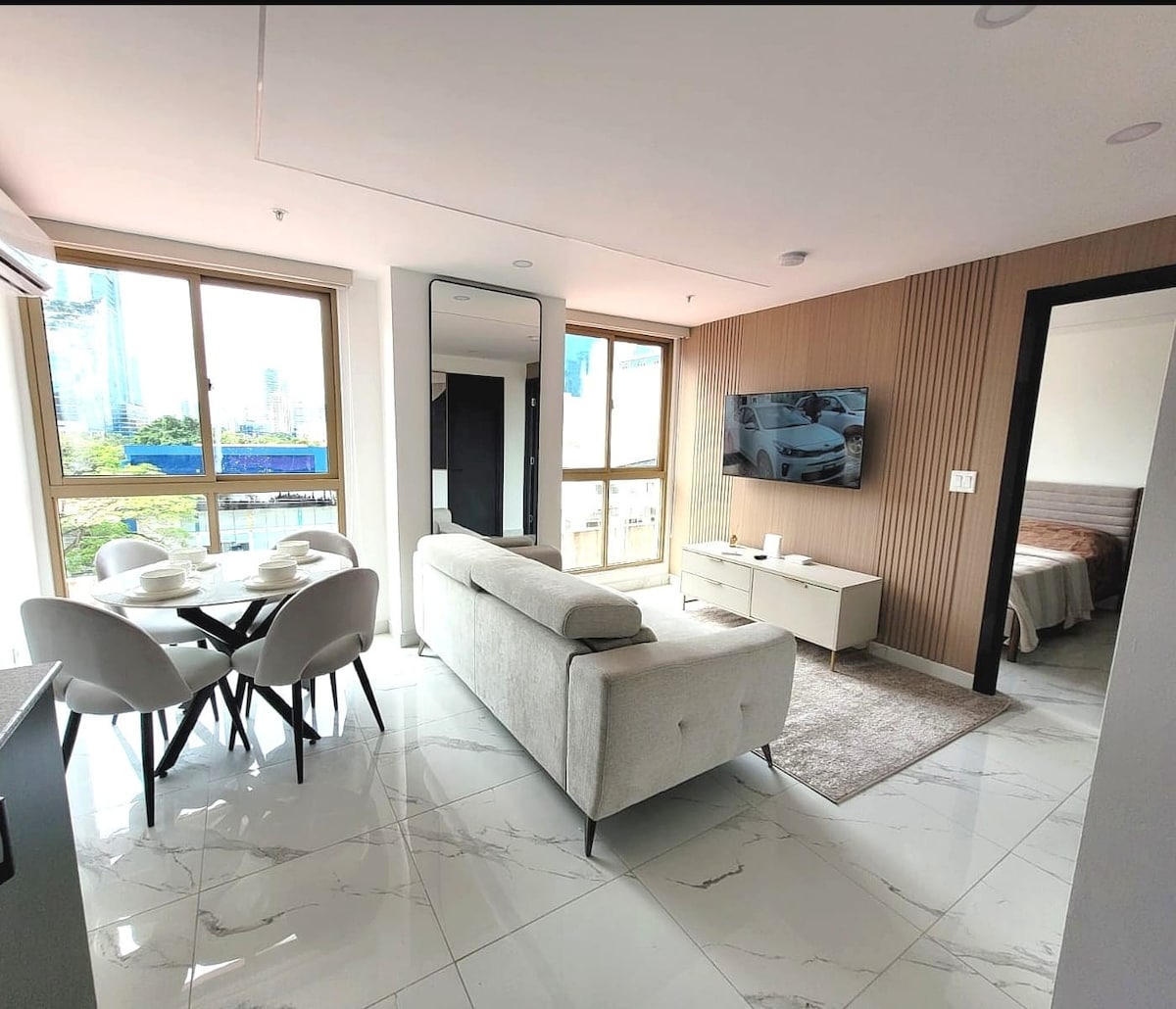 A modern living space features a light-colored sofa facing a wall-mounted television. A round dining table with four chairs is positioned nearby. Large windows offer views of the cityscape, while natural light brightens the room. The elegant flooring complements the contemporary design.