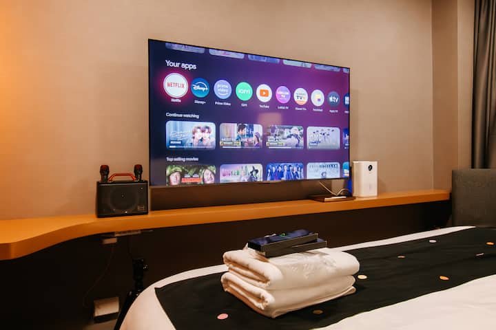 Modern bedroom with a large smart TV offering streaming apps, a sleek console shelf, and compact speaker. Fresh towels are neatly placed on the bed, adding a hotel-style touch to this cozy, well-equipped space.