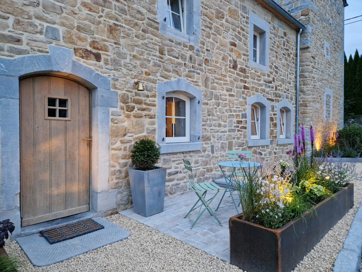 The exterior features a charming stone facade, complemented by a wooden door with a small window. A patio area is highlighted by a small table and green chairs, surrounded by vibrant flowers and plants, illuminated by soft outdoor lighting.