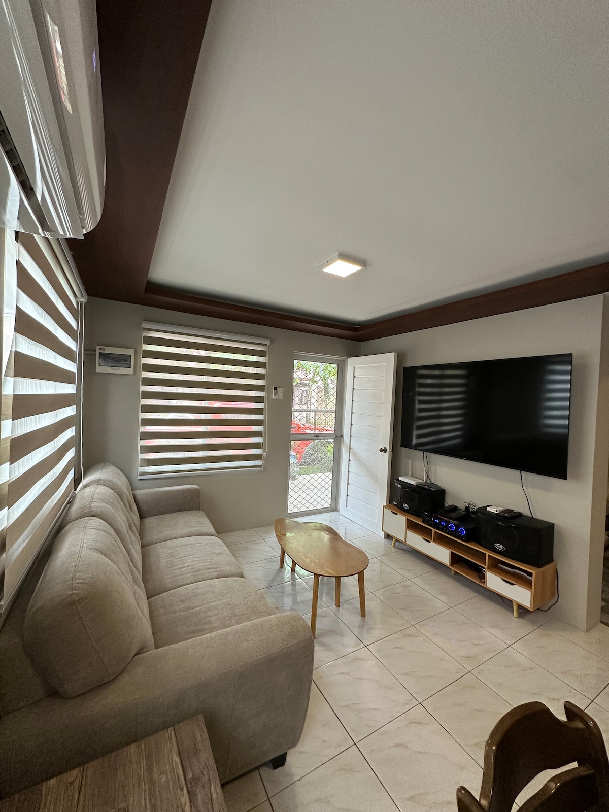 A comfortable living area features a light-colored sofa and a wooden coffee table. A large flat-screen TV is mounted on the wall, and sunlight filters through striped window shades. An inviting atmosphere is created by the neutral-toned walls and tiled flooring.