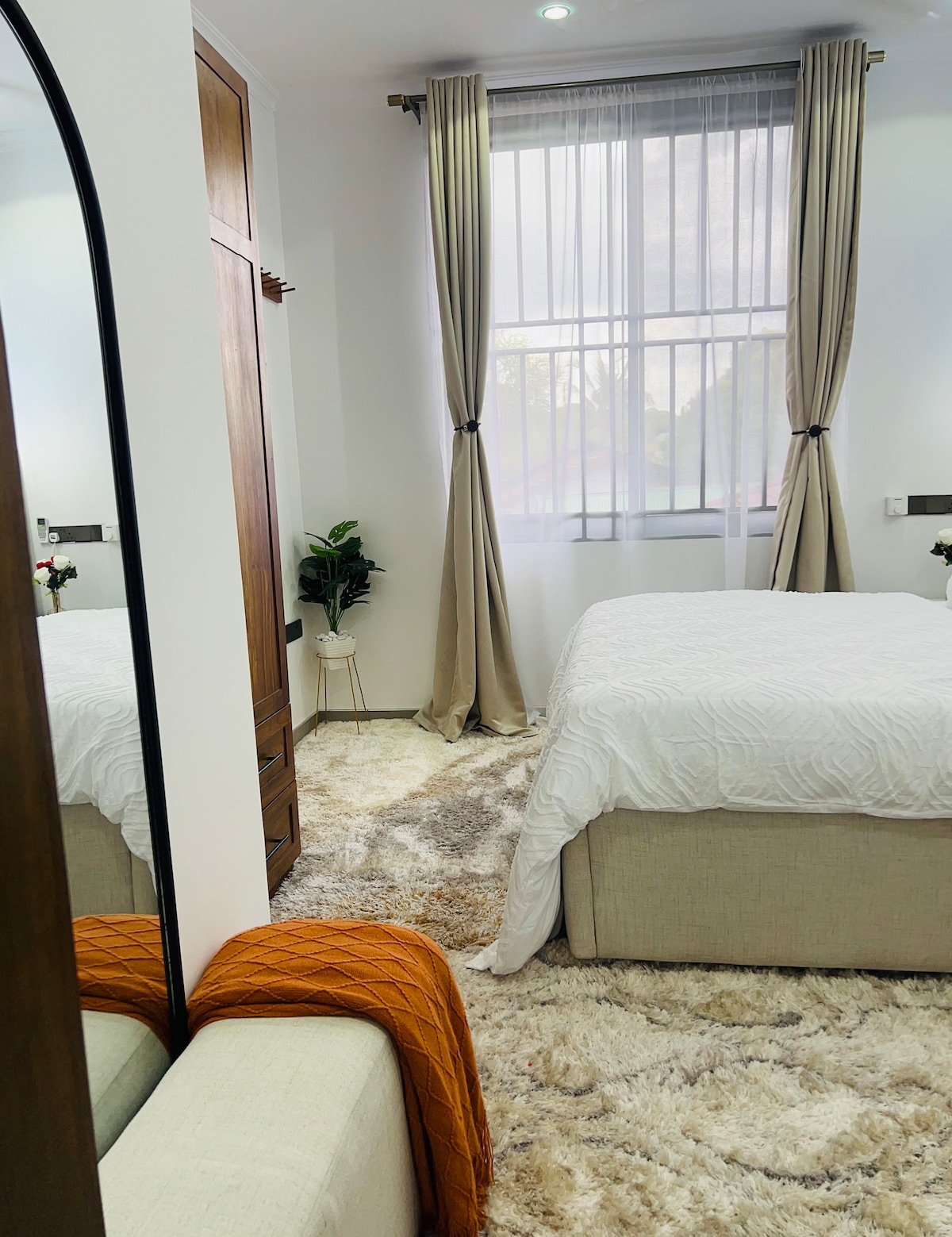 A comfortable bedroom is presented with a spacious bed covered in a white bedspread. Soft area rugs cover the floor, while natural light filters through sheer curtains. A mirror reflects a cozy chair and decorative plant, adding a touch of green to the warm space.