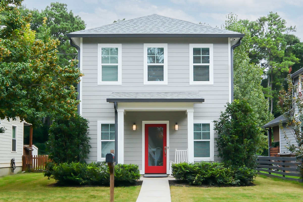 The exterior of the home features a light gray facade with white trim and a welcoming red front door. Lush green landscaping surrounds the walkway leading to the entrance, and large windows invite natural light into the space. The two-story structure is complemented by a charming porch.