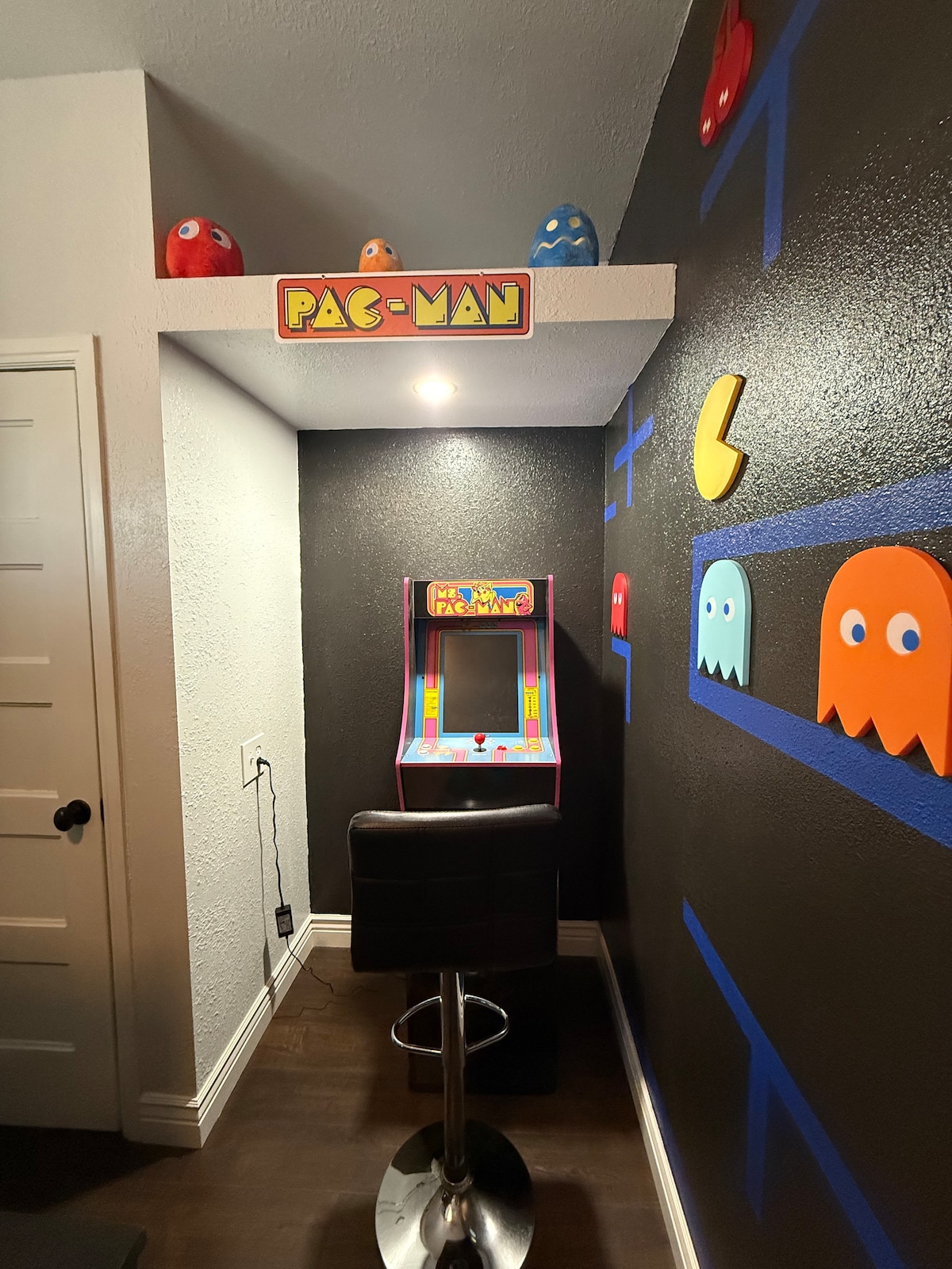 A vibrant game room features a classic Pac-Man arcade machine positioned against a dark wall. Colorful ghosts and a yellow Pac-Man character are painted on the wall above and around the machine. A bar stool is placed next to the machine, creating a playful space for enjoyment.