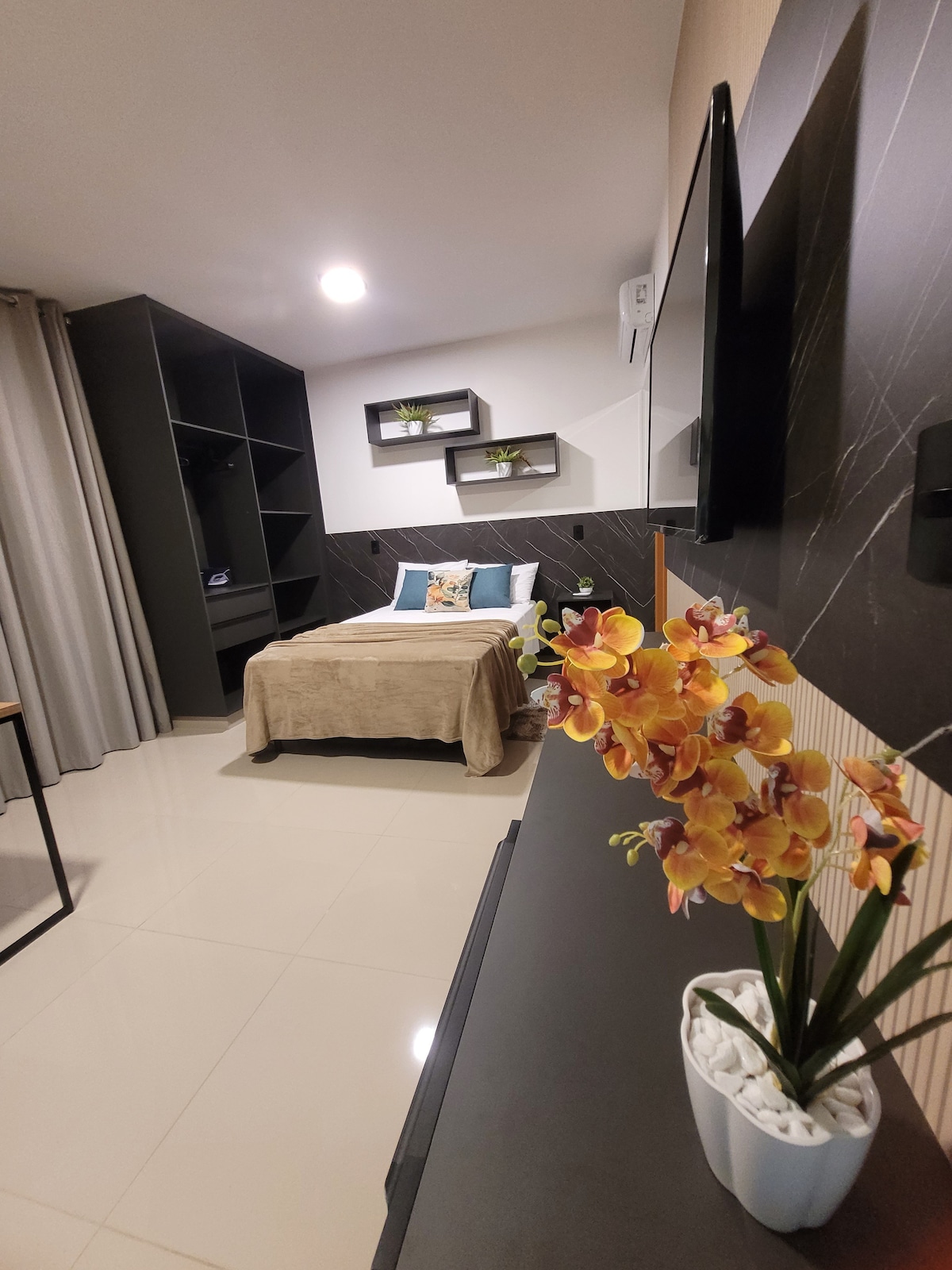 A modern sleeping area features a cozy bed with a light-colored coverlet, complemented by decorative pillows. A wall-mounted television is visible above a sleek surface, while shelves with greenery provide subtle accents. Natural light enters through soft curtains, enhancing the inviting atmosphere.