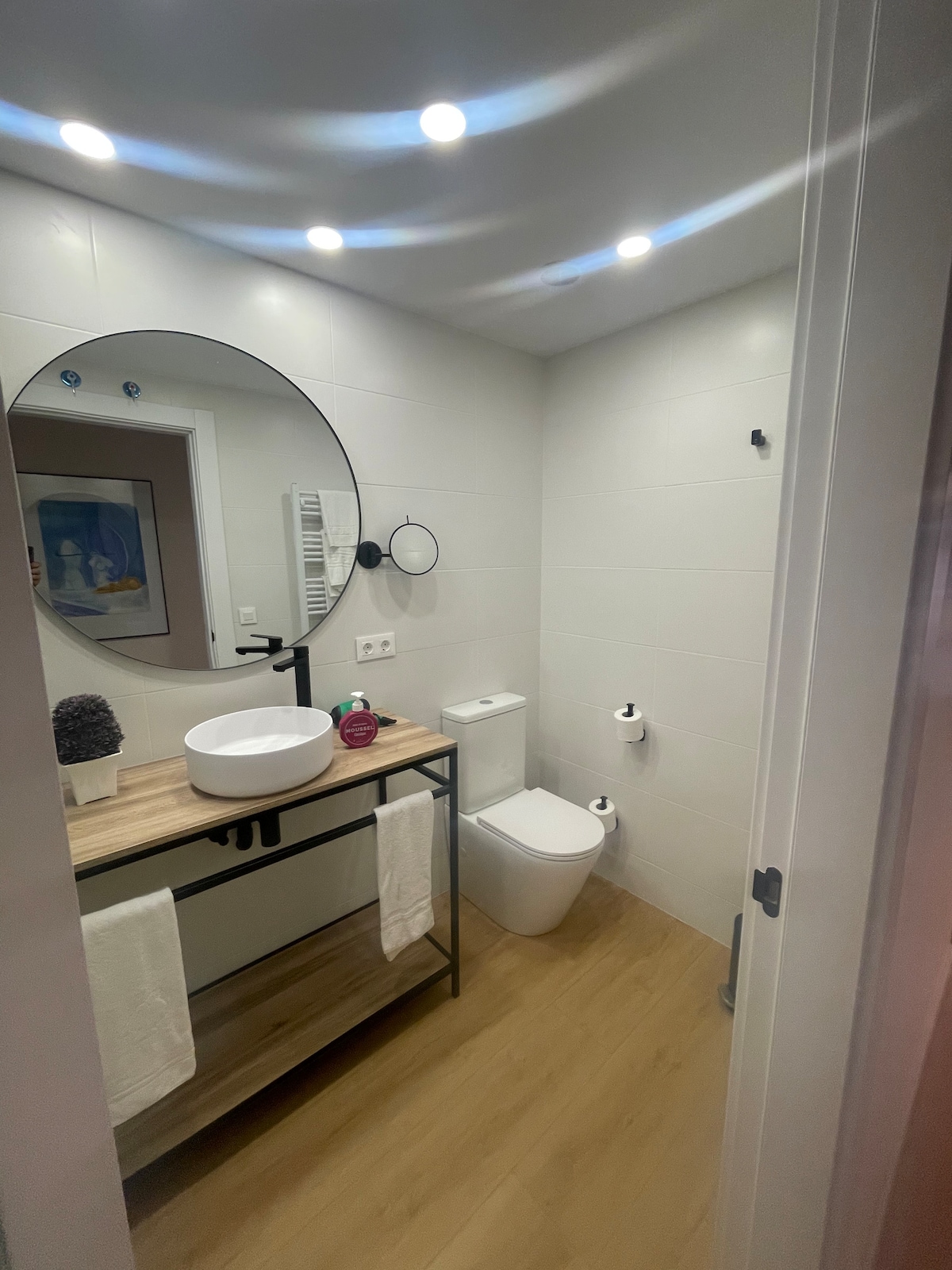 A modern bathroom is presented with neutral-toned tiles and warm wood flooring. A round mirror is mounted above a wooden sink console that features a white basin. Essential amenities are neatly arranged, including towels. Soft lighting enhances the room's clean and serene appearance.