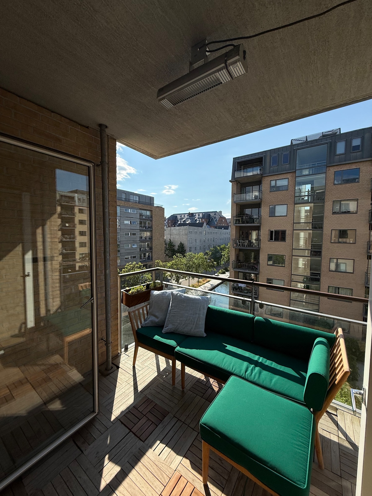 A private balcony features a green seating area with a comfortable sofa and matching ottoman. Natural light filters through, enhancing the view of surrounding buildings and greenery. Wooden flooring and potted plants complete the inviting outdoor space.