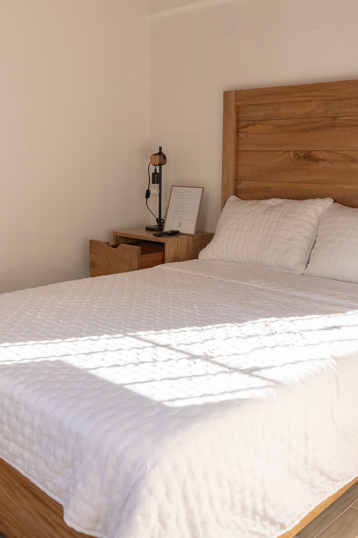 A cozy bedroom features a wooden bed frame with a neatly made white quilt. A bedside table holds a lamp and a framed document, while soft natural light filters through a nearby window, casting gentle shadows across the room.