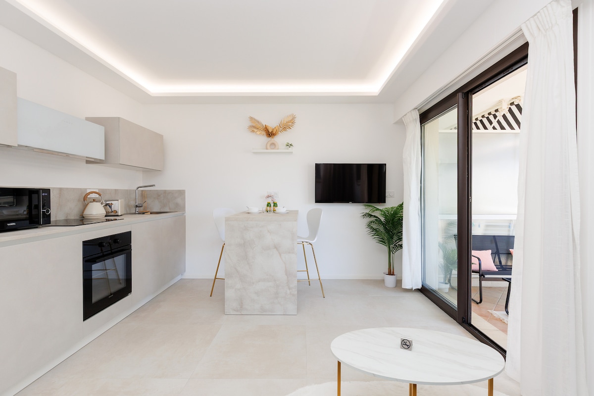A modern, minimalist kitchen and living area is presented, featuring light-toned cabinetry and integrated appliances. A small island with seating is positioned under soft lighting. A wall-mounted TV is visible, with large glass doors leading to the outdoor terrace, allowing natural light to fill the space.