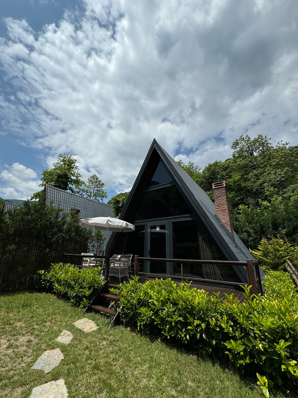 The exterior of a modern A-frame house is captured under a partly cloudy sky. A shaded seating area is visible, featuring a small table and chairs. Lush greenery surrounds the property, and a stone pathway leads to the entrance, enhancing the serene outdoor setting.