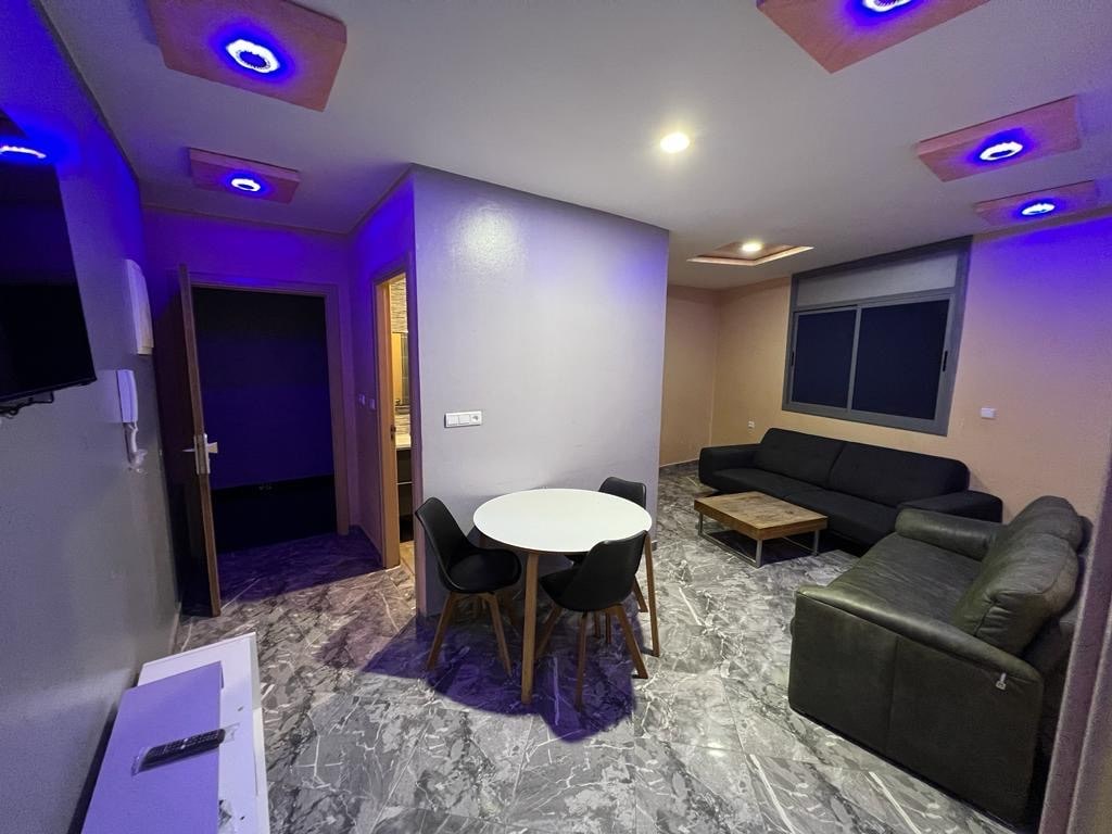 A modern living area features a circular dining table surrounded by black chairs. A dark sofa is positioned adjacent to a light-colored coffee table. Soft lighting emanates from the ceiling, creating a calming ambiance against the contrasting wall colors.