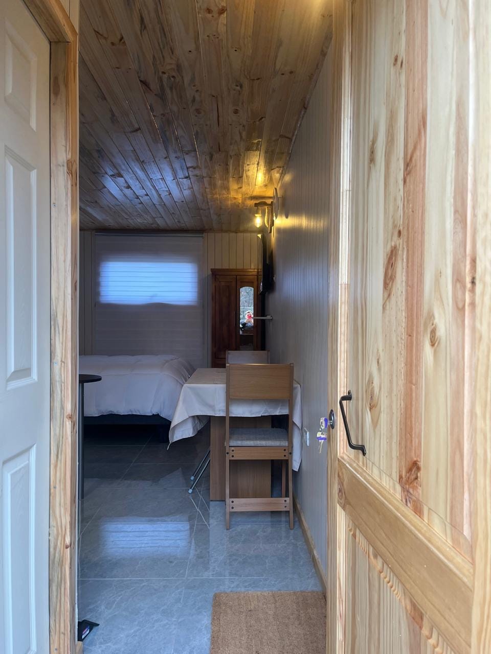 A doorway opens to a cozy interior space featuring wooden walls and ceiling. A spacious bed is visible in the distance, alongside a small table and chair. Natural light enters through a window, illuminating the polished floor tiles.