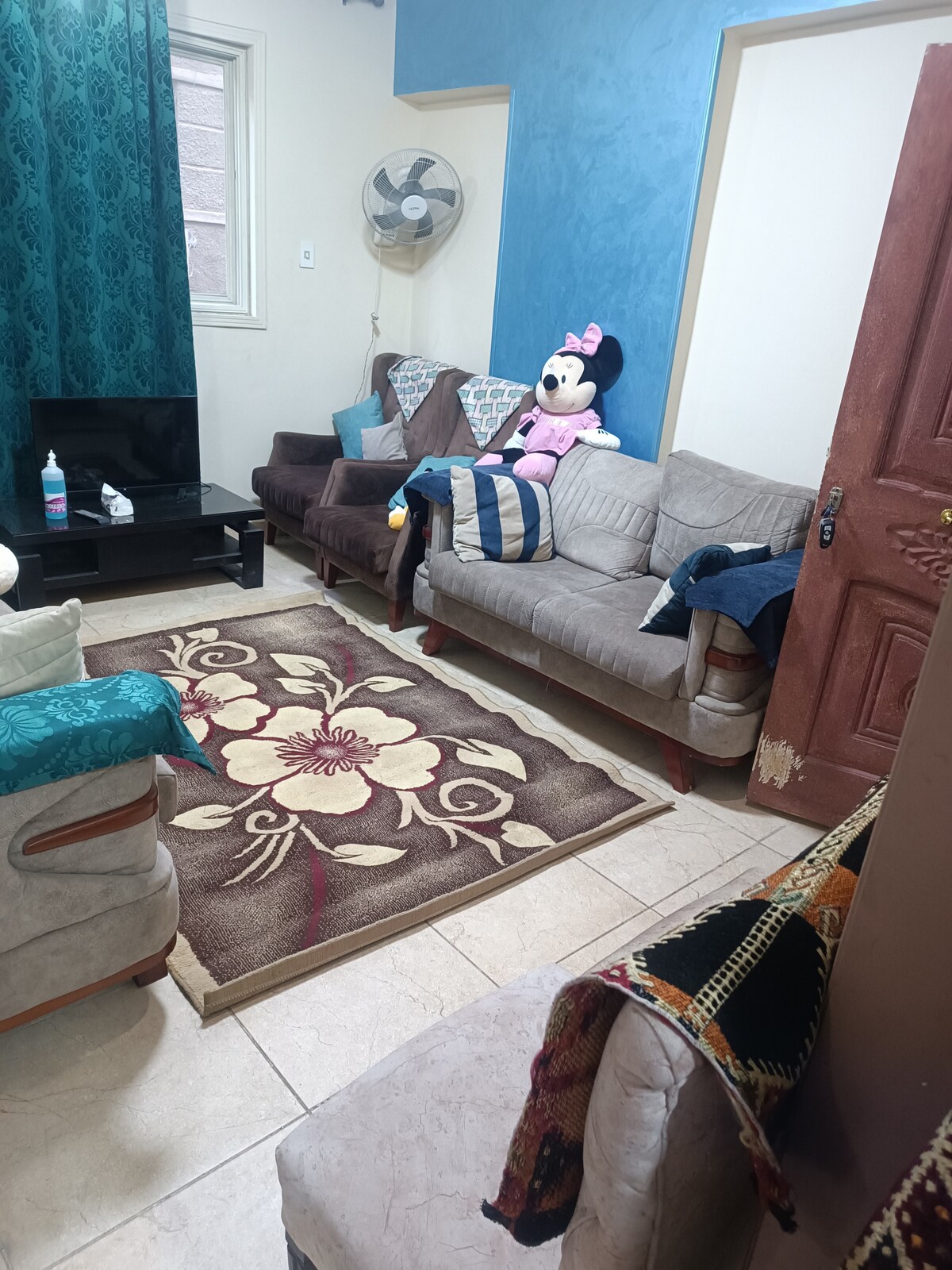 A cozy living area features a central area rug adorned with a floral design. The space contains several armchairs upholstered in various fabrics, with a small television positioned on a low table. A fan is mounted on the wall, and a plush toy is seated on one of the chairs.