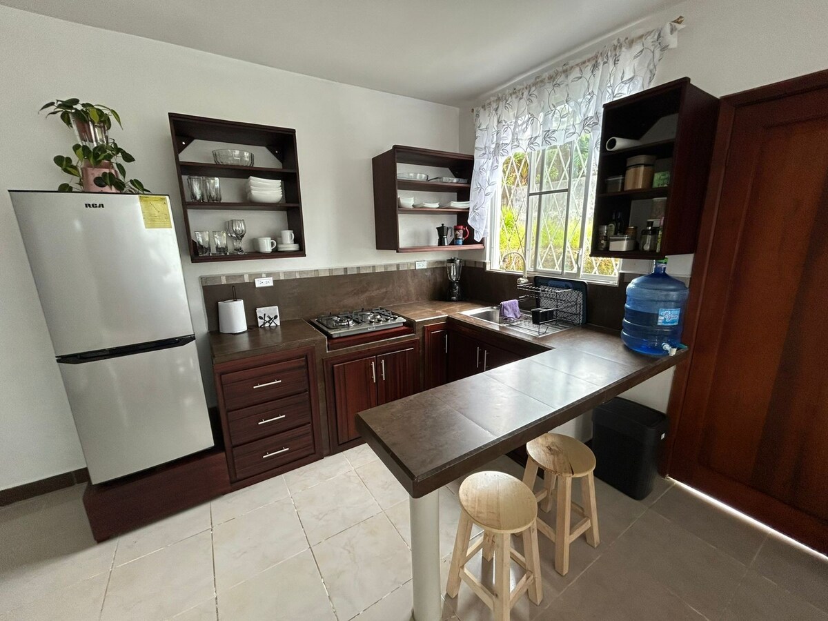 The kitchen features dark wooden cabinetry and open shelves displaying various dishware. A glass-top stove and a stainless steel refrigerator are included. A small dining area with two wooden stools is positioned near a window, which is dressed with light, sheer curtains.