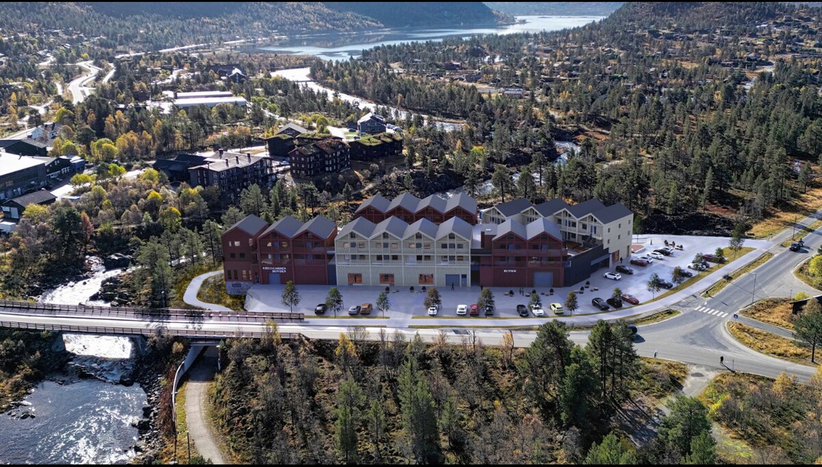 An aerial view of a modern building complex surrounded by natural landscapes. The structure is positioned near a river with a bridge, showcasing greenery and mountain scenery in the background. Parking spaces are visible alongside the building, indicating convenient access.