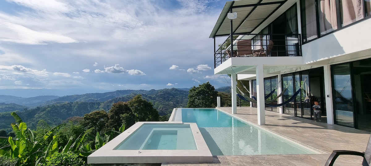 An expansive outdoor area features a sleek infinity pool and a separate jacuzzi, surrounded by lush greenery and rolling hills. Comfortable hammocks hang between sturdy posts, with large glass doors leading into the villa, offering uninterrupted views of the scenic landscape.