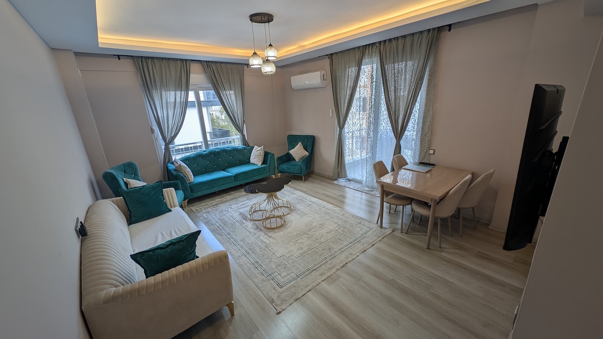 A spacious living room is displayed, featuring comfortable seating arrangements with teal and cream-colored sofas. A round coffee table sits in the center, complemented by a dining table nearby. Large windows allow natural light to fill the space, while soft curtains enhance the ambiance.