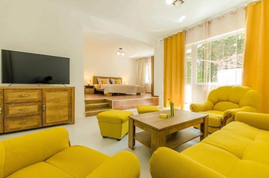 The living area features a pair of yellow armchairs and a matching sofa surrounding a wooden coffee table. A television is mounted on the wall opposite. Sunlight streams through large windows dressed in sheer curtains, illuminating the room and leading to a visible bedroom area.