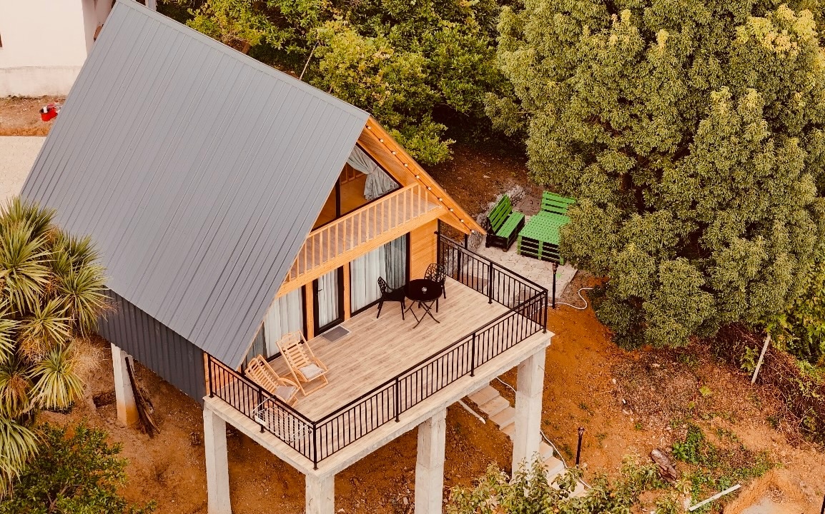 A modern cabin is elevated on sturdy pillars, surrounded by lush greenery. A spacious deck features a circular table and two chairs, inviting relaxation. The sloped roof, complemented by wooden accents, adds a contemporary touch to the serene outdoor setting.