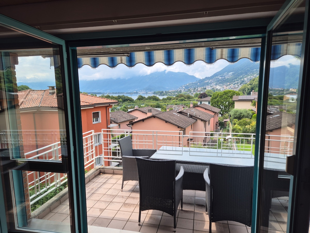 A private terrace is presented with a dining table and chairs, framed by glass doors. Scenic views of the surrounding hills and lake are visible, offering a tranquil outdoor space for relaxation.