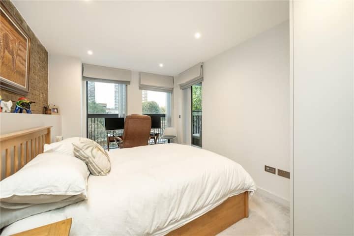 Chumba cha kulala chenye mwanga na kitanda cha watu wawili, mashuka laini, dawati la kazi karibu na madirisha makubwa na mwonekano wa kijani cha jiji.