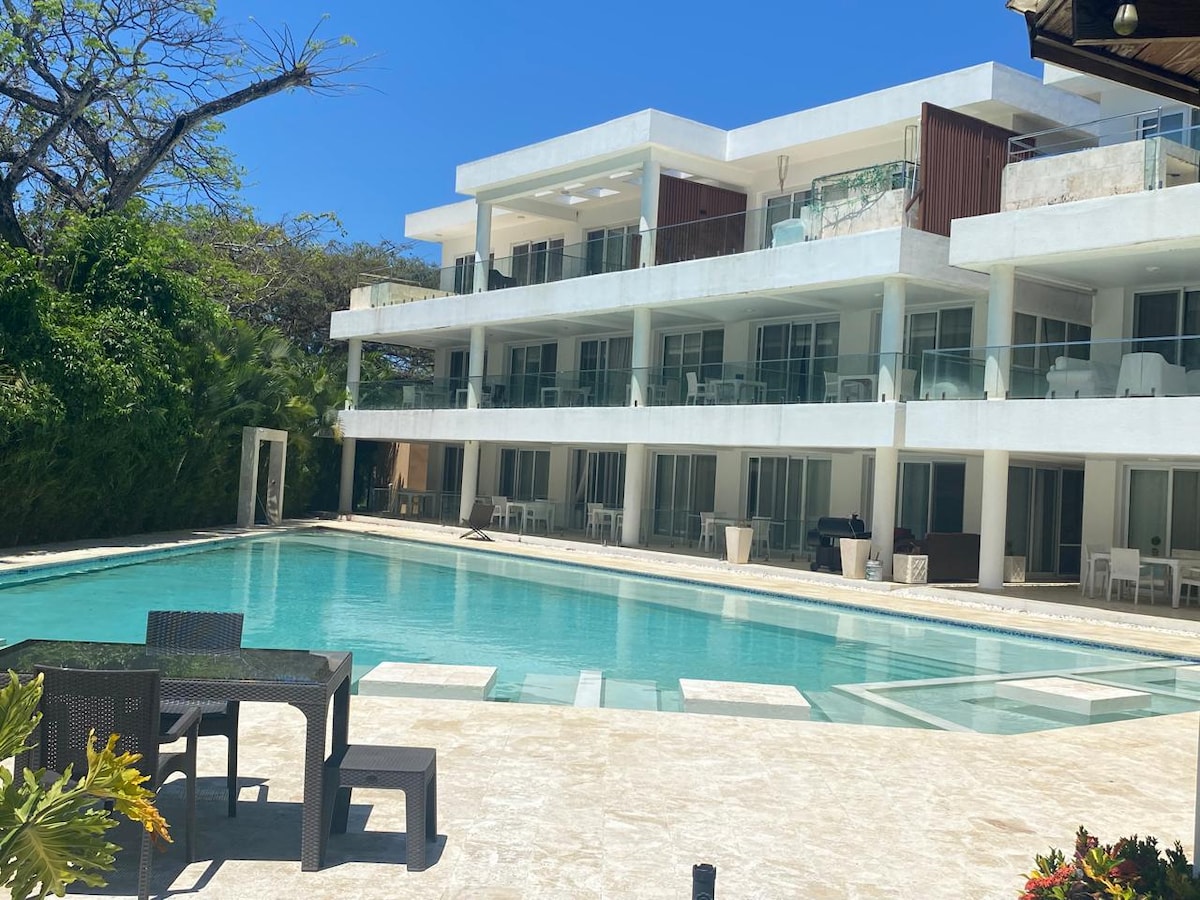 A modern building with multiple levels surrounds a rectangular swimming pool. Lounge areas are visible, along with outdoor seating and tables. Lush greenery is present in the background, complementing the clear blue sky.