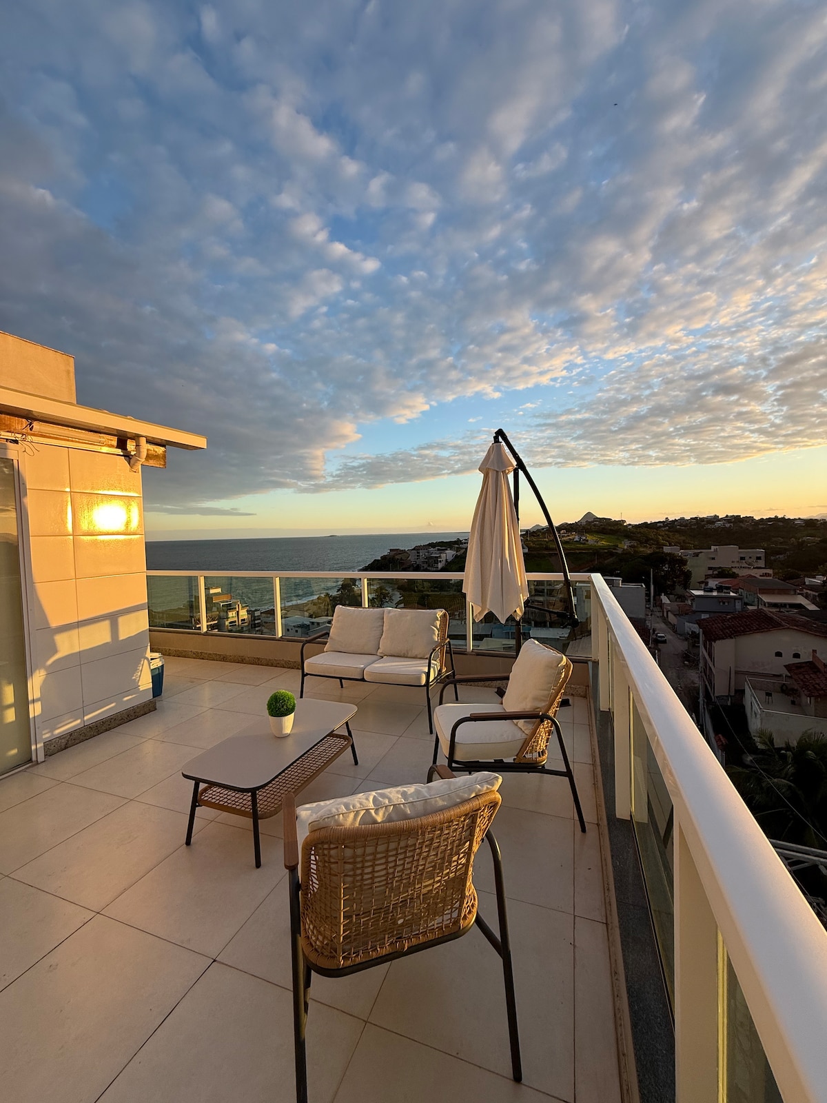 An outdoor terrace is presented with seating made up of two cushioned chairs and a small table, all positioned to overlook the ocean. A large umbrella is also visible, providing shade. The sky displays a mixture of clouds with a warm sunset glow.