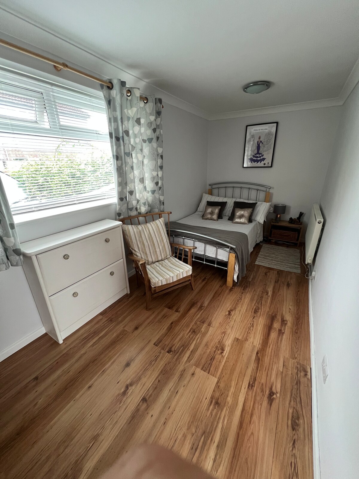 A bright and tidy double bedroom features a small double bed with bedding and two decorative pillows. A wardrobe and a chest of drawers provide storage, while a wooden chair is positioned beside the window dressed with light grey curtains. Natural light fills the room.