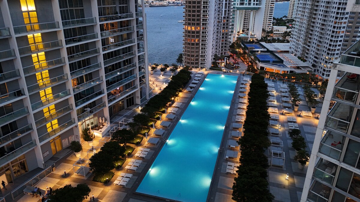 An aerial view of a well-lit pool area is presented, showcasing a long, rectangular pool surrounded by lounge chairs and landscaped greenery. Nearby buildings and pathways are illuminated, creating a lively ambiance at night, with the shimmering water reflecting nearby lights.