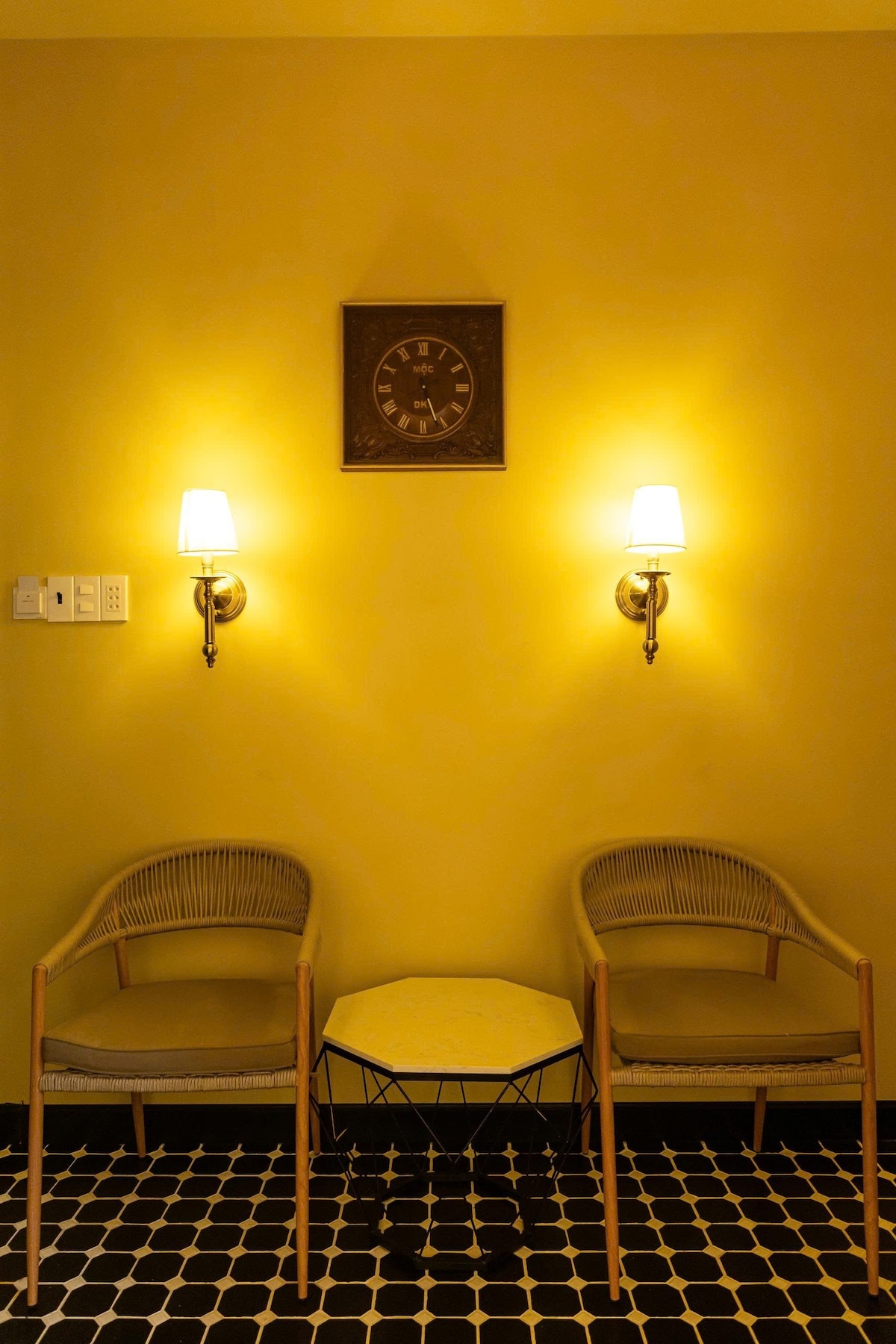 A cozy seating area is featured with two rattan chairs positioned beside a small, round table. The warm yellow walls are illuminated by soft wall sconces, and a clock is mounted above the chairs, contributing to a relaxed ambiance.