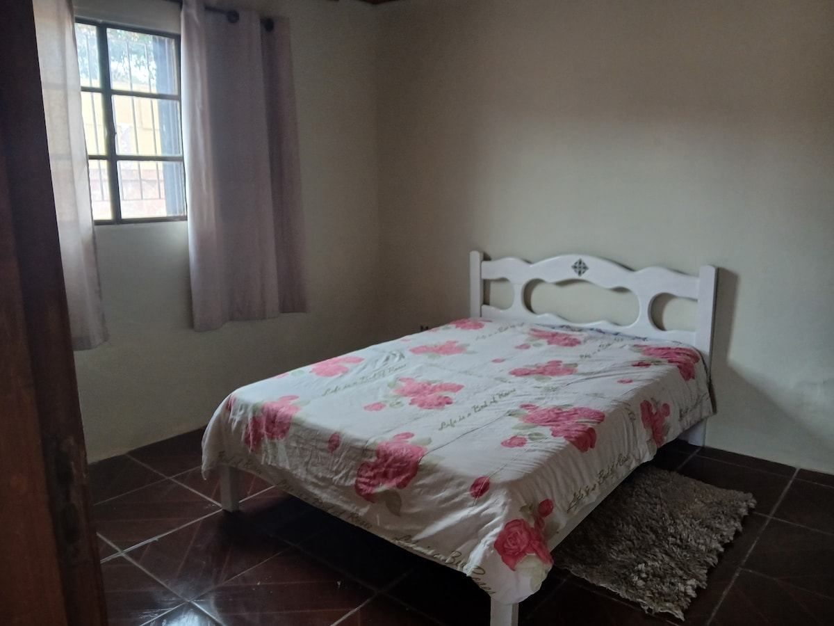 A simple bedroom features a spacious bed with a floral-patterned coverlet, complemented by soft curtains framing a nearby window. The floor is tiled, and a small decorative rug is placed beside the bed, creating a cozy atmosphere in the room.