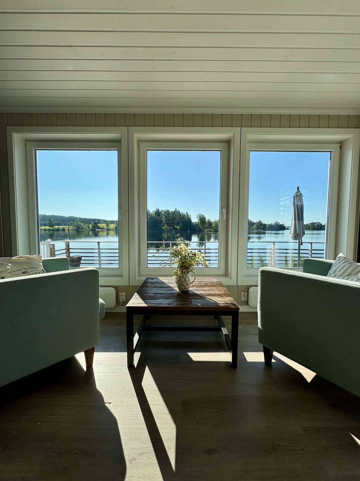 Large windows frame views of Einafjorden and the surrounding landscape. A wooden coffee table sits between two soft-green armchairs, inviting moments of relaxation. The natural light illuminates the space, enhancing the serene atmosphere created by the water and greenery beyond.