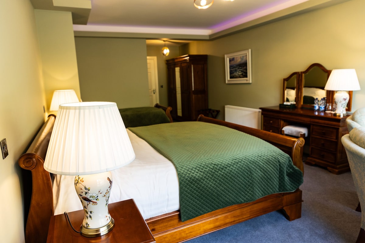 A spacious bedroom features a wooden sleigh bed dressed in crisp white linens and a green quilt. Soft lighting is provided by bedside lamps, and a wooden dresser and mirror create a cozy atmosphere. A wardrobe is also present, adding to the room's functionality.
