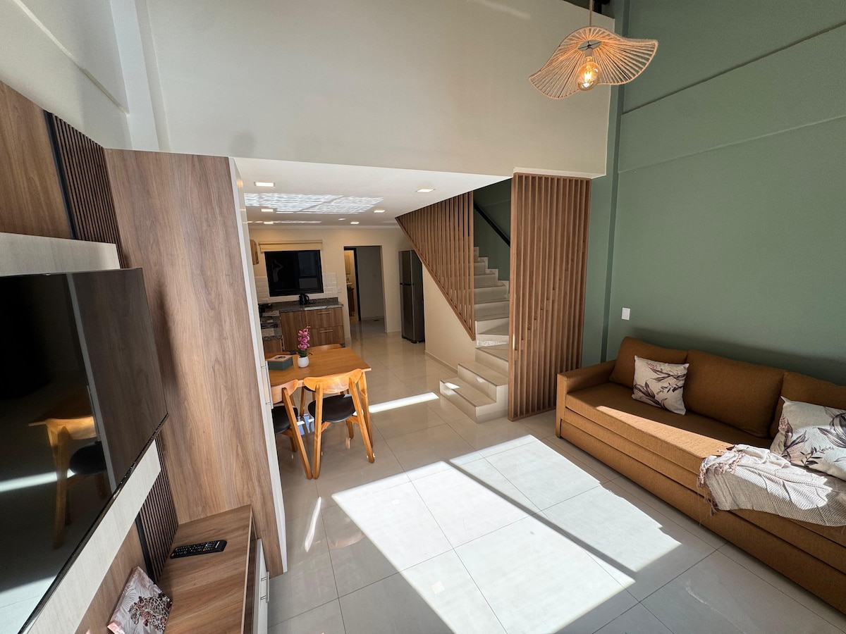 A modern living area features a warm-colored sofa facing a wall-mounted television. A wooden dining table is positioned near large windows that allow natural light to illuminate the room. Stairs rise to a second level, and light greets the space through a decorative pendant light overhead.