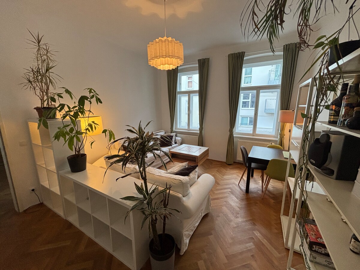 A living area features a mix of greenery and modern furnishings. A white sectional sofa and wooden coffee table are complemented by floor lamps and indoor plants. Large windows allow natural light to fill the space, while a dining table is seen in the corner near a bookshelf.