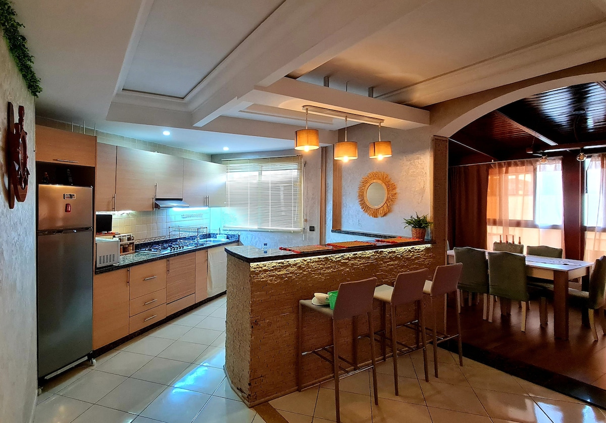 A modern kitchen area features sleek cabinetry, stainless steel appliances, and a spacious countertop that separates it from a dining area. Bar stools are situated at the counter, while natural light filters through the windows, highlighting the dining table with upholstered chairs in the adjoining space.