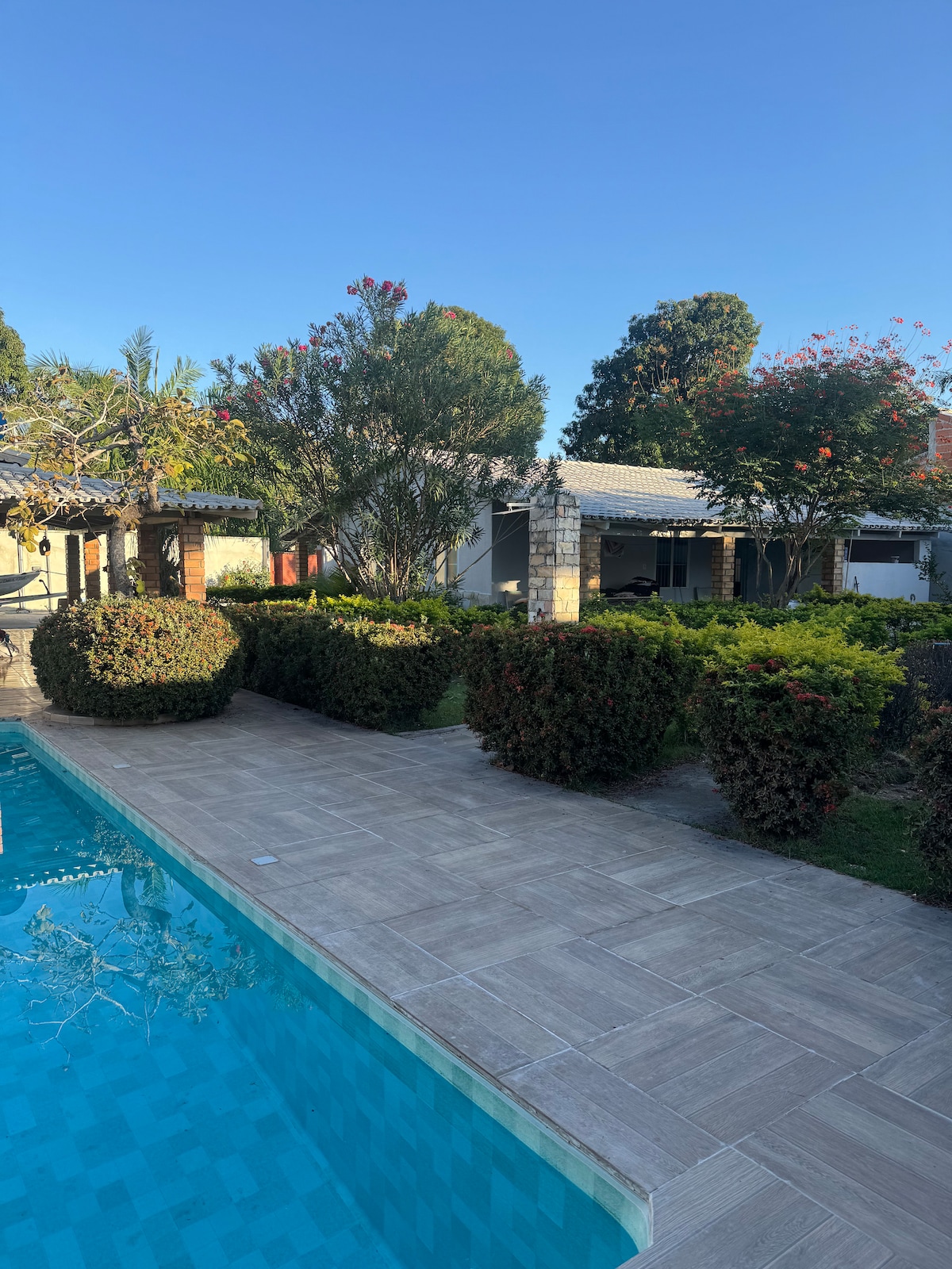 A clear view of the property showcases a glistening pool surrounded by well-maintained gardens. Lush greenery and flowering bushes provide an inviting atmosphere, while the main house features a covered patio area visible in the background, perfect for enjoying sunny days.