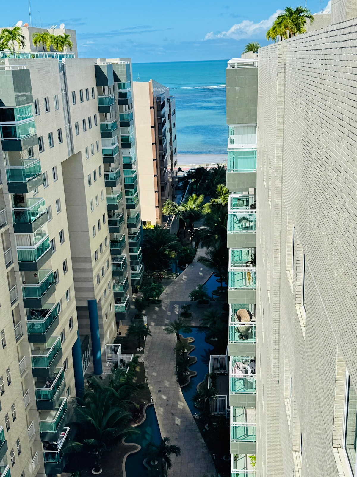 A view of the boulevard and ocean is presented between two tall buildings. A landscaped pathway lined with tropical plants is visible, leading toward the water. The clear sky and distant ocean enhance the visual appeal of the surrounding architecture.