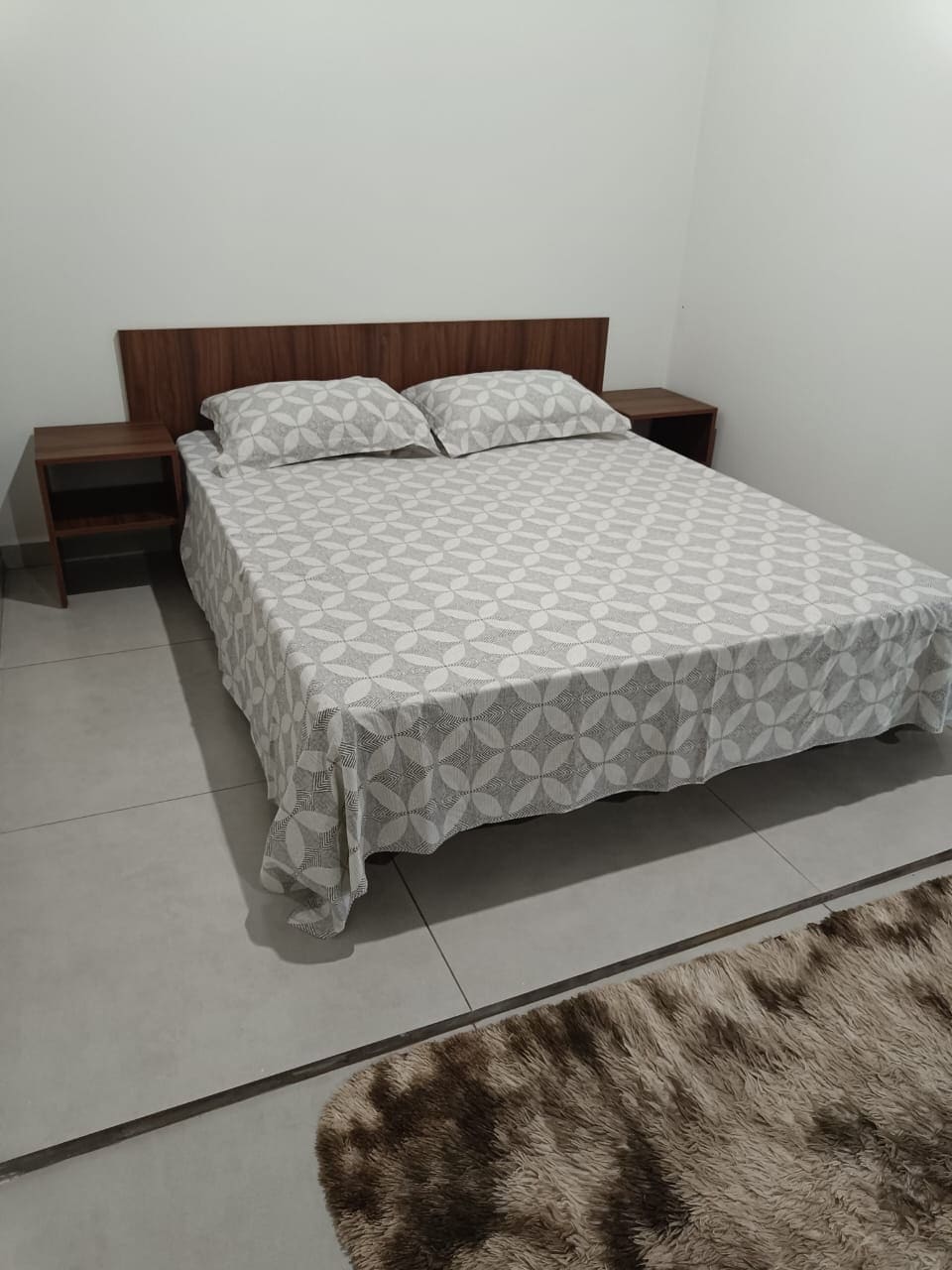 A compact bedroom is presented, featuring a queen bed with a patterned bedspread. Two nightstands are positioned on either side of the bed, while a plush area rug lies at the foot, contributing to a comfortable and functional space.