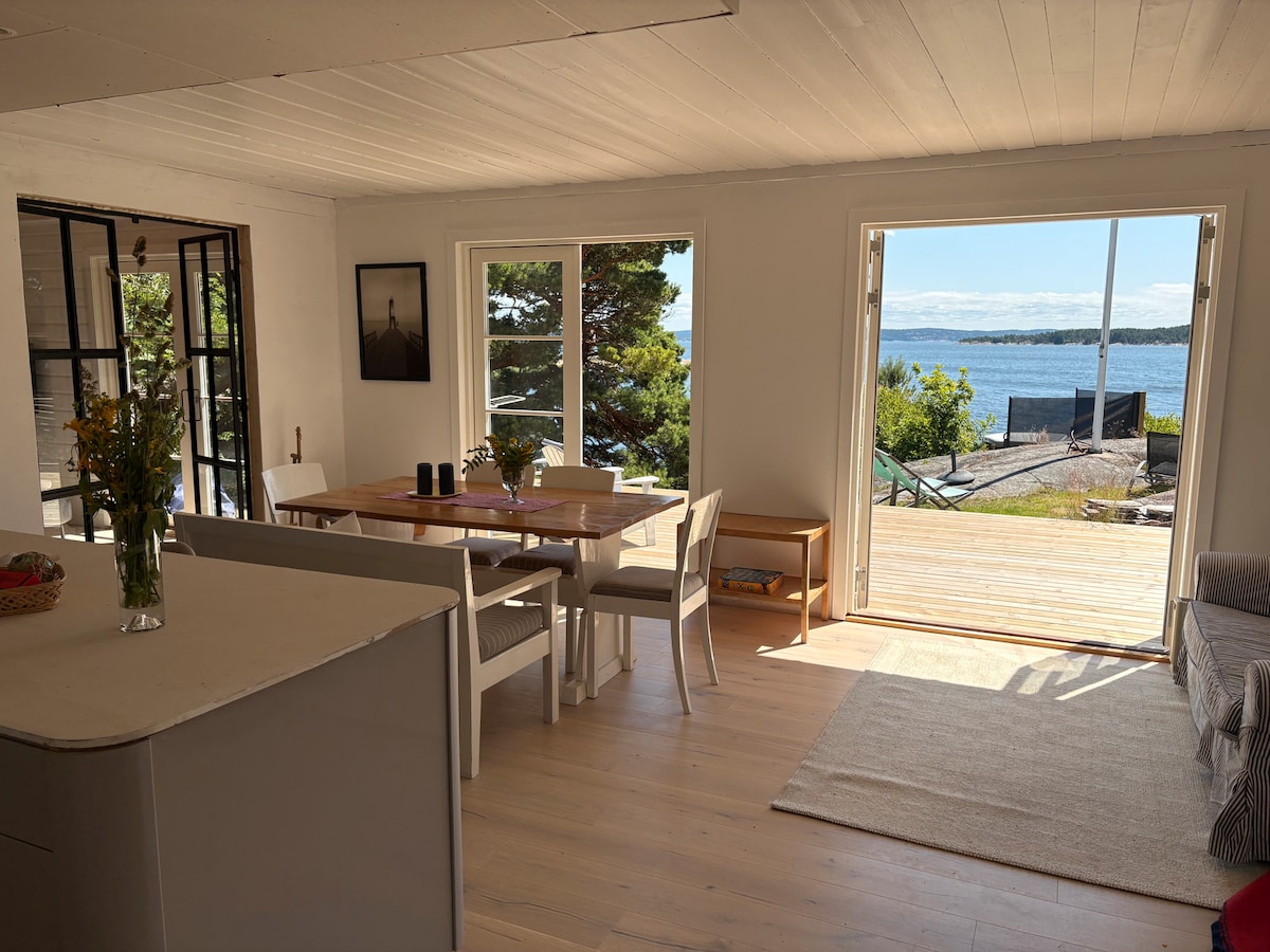 A bright living space is featured, with large glass doors opening to a view of the water. A wooden dining table with several chairs occupies the center, while a cozy seating area with a sofa is visible to one side. Fresh flowers on the table add a natural touch.