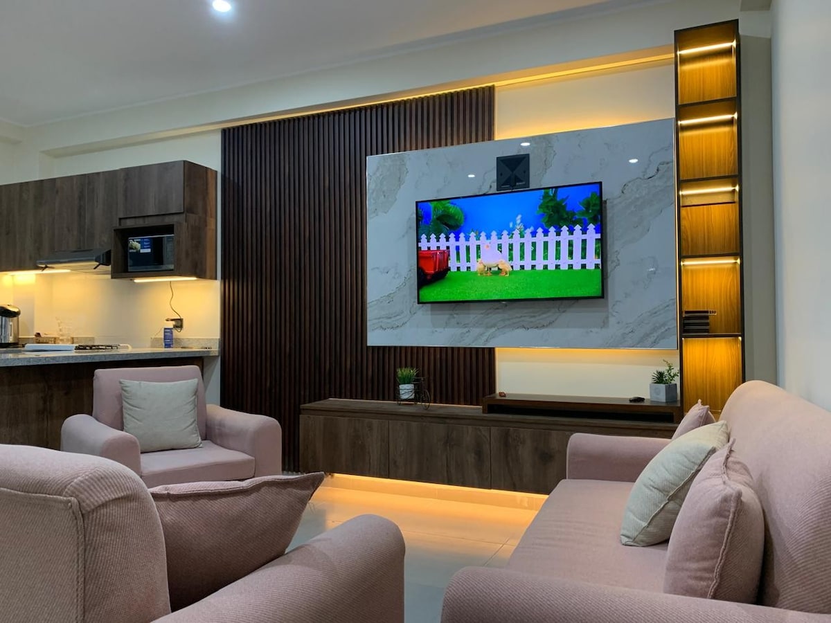A modern living area is presented, featuring soft-toned sofas arranged around a stylish wall-mounted TV. The wall is adorned with a marble finish and wooden accents, providing a warm atmosphere, while the space is enhanced by subtle LED lighting.