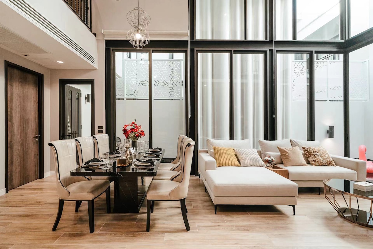 A modern and elegantly designed living and dining area is featured, with a dark dining table set for six, complemented by beige, upholstered chairs. A light-colored sectional sofa sits nearby, accented with a decorative pillow. Large glass windows allow natural light to fill the space.