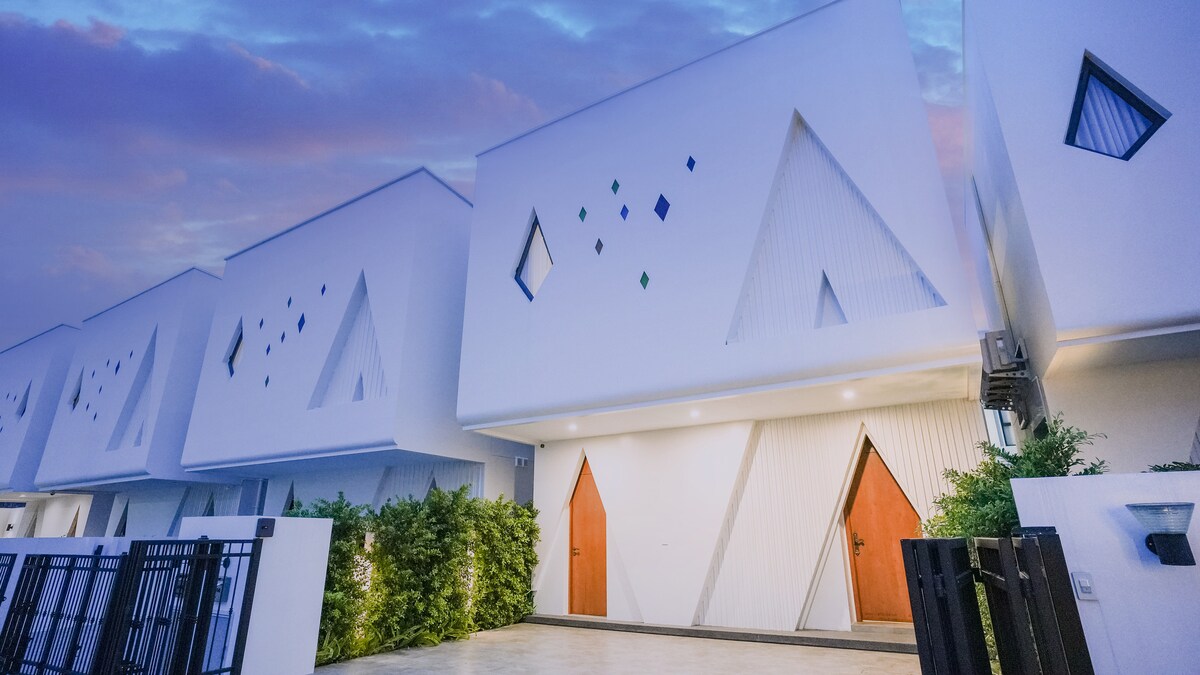 A modern villa exterior showcases a striking design with angular geometric shapes and vibrant colored accents. The front entrance features two distinct wooden doors, framed by lush greenery. Soft evening light enhances the clean lines of the architectural structure against a backdrop of a twilight sky.