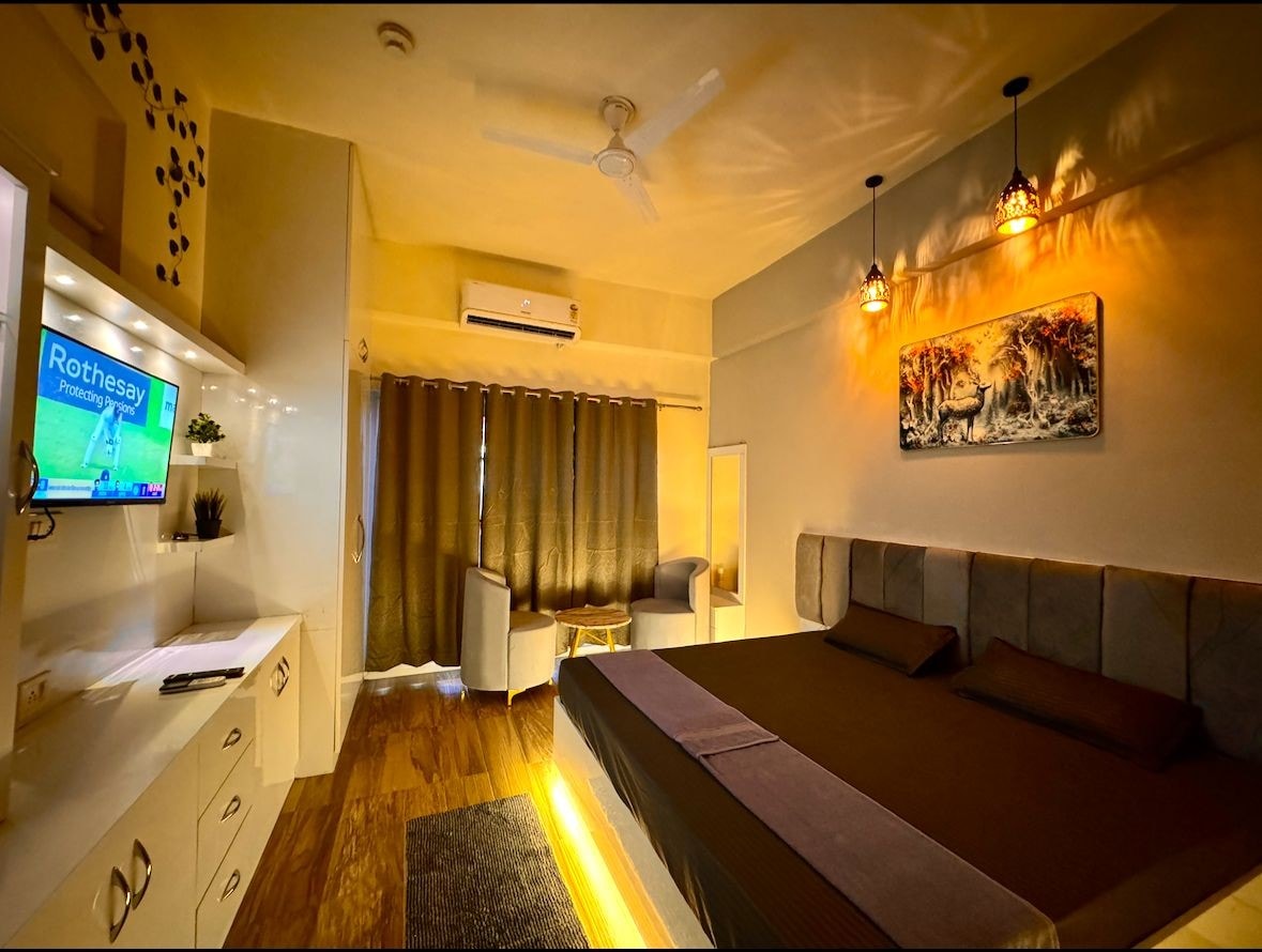 A spacious sleeping area is highlighted by a large bed with a dark bedspread. Soft lighting emerges from two pendant lights above, and a colorful artwork adorns the wall. A modern TV and a minimalist white cabinet are present, complemented by a sitting area with two chairs near a window with dark curtains.