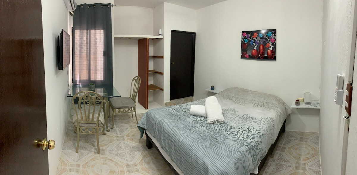 A well-lit room features a spacious bed with a light grey patterned blanket. Two wooden chairs are positioned near a small glass table. Shelving is present against one wall, while a colorful piece of wall art adds a pop of color. Natural light filters in through the window.