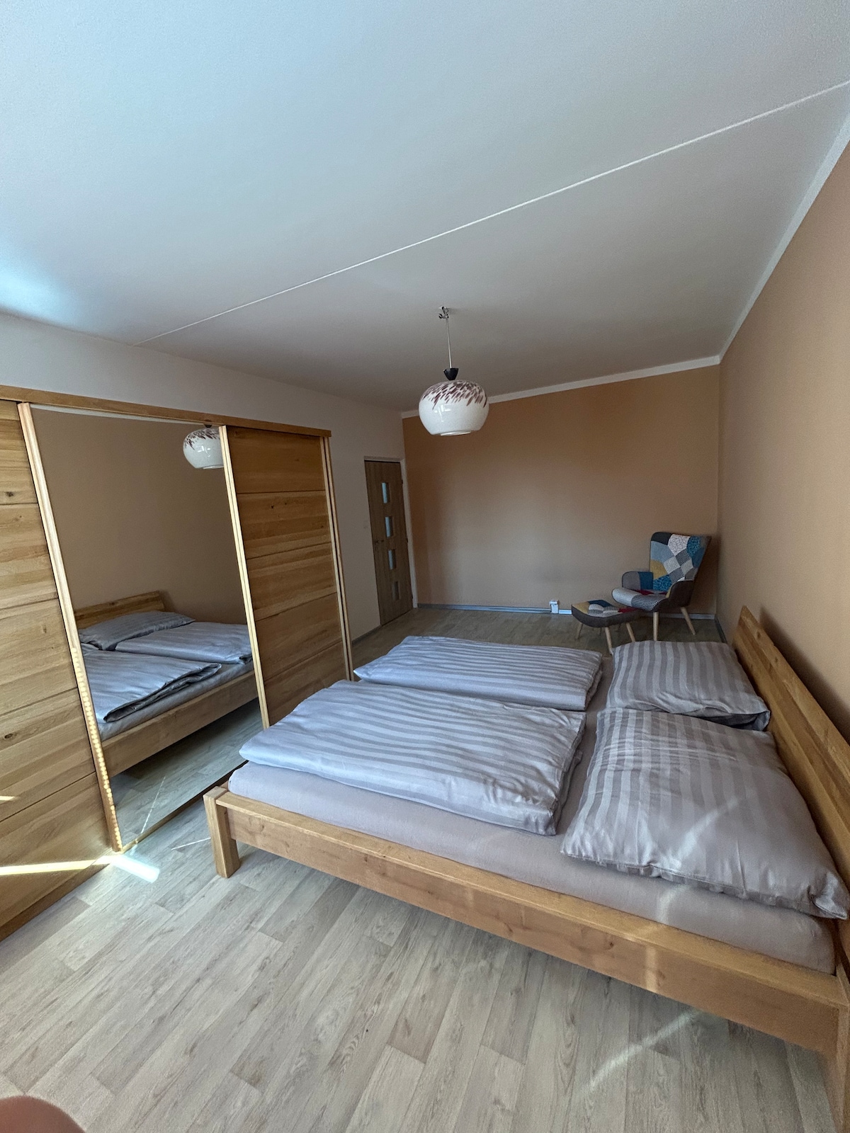 A spacious bedroom is illuminated by natural light filtering through a window. Two wooden beds are dressed with soft striped bedding. A wardrobe with mirrored doors reflects the room, and a comfortable armchair adds a cozy touch, enhancing the room's functionality.