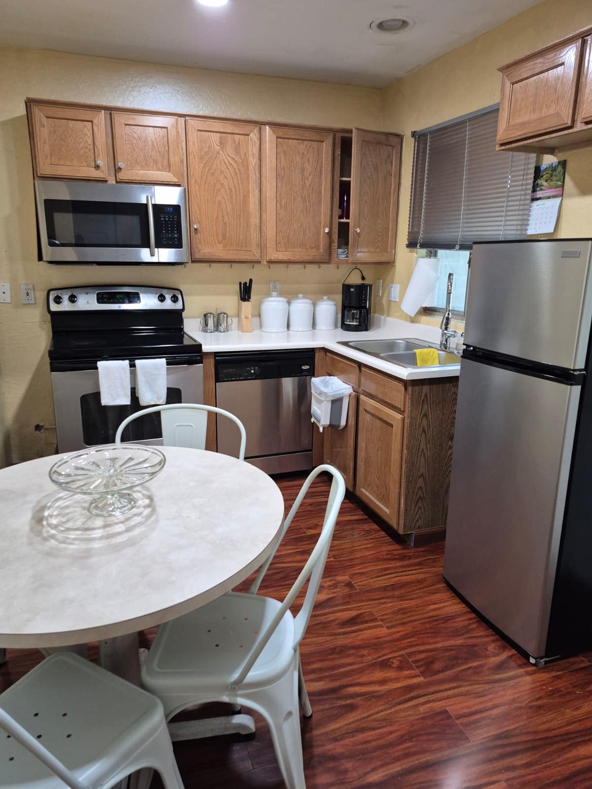 A compact kitchen area features wooden cabinetry and modern appliances. A round table with two chairs is positioned beside the counter, promoting a functional dining space. White dishware and utensils are neatly arranged, while soft light enters through the window, enhancing the warm tones of the wood.