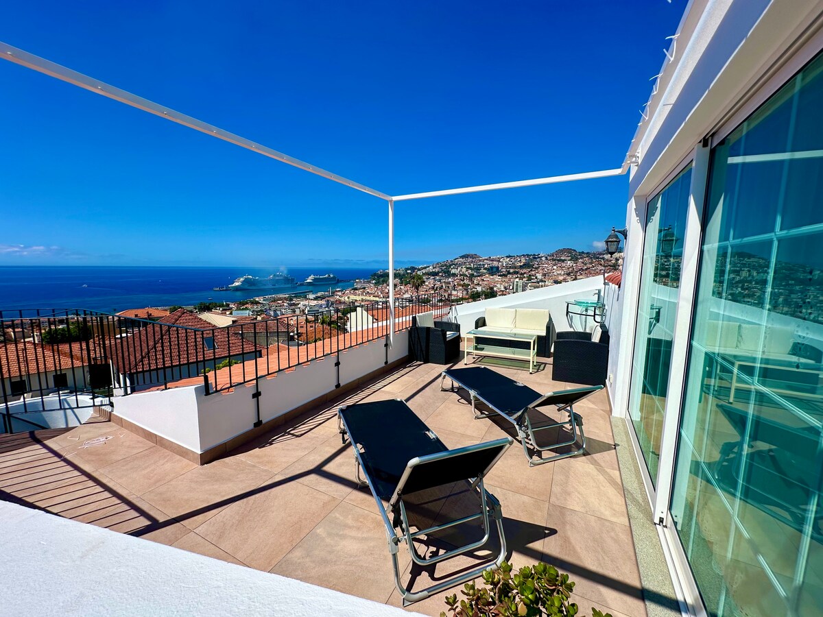 The terrace offers stunning panoramic views of the sea and city, featuring several lounge chairs positioned for relaxation. The space is surrounded by low walls, enhancing the openness while providing a comfortable atmosphere during sunny days.